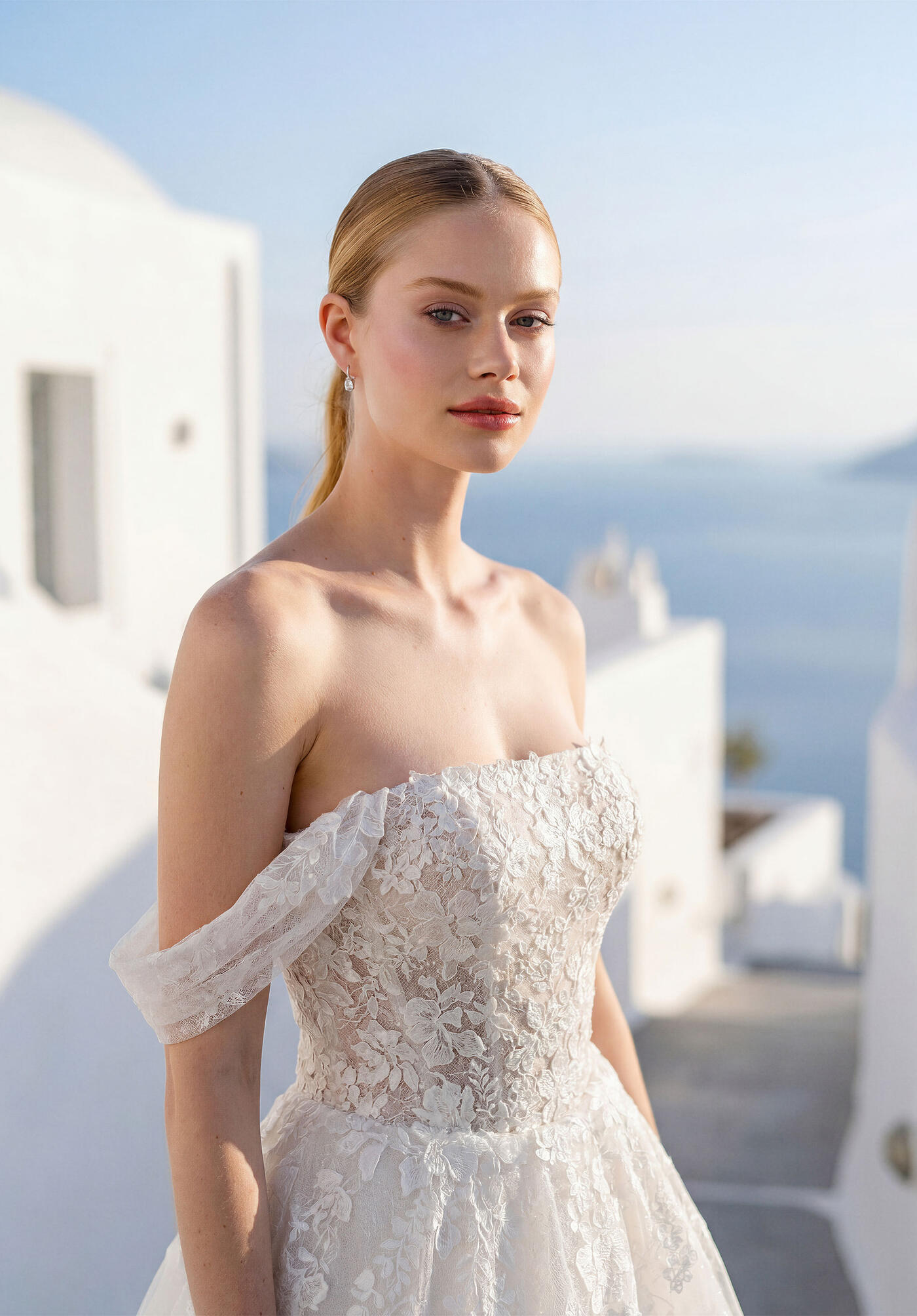 A bride in a Morilee floral lace gown with off-the-shoulder sleeves, the sea behind her
