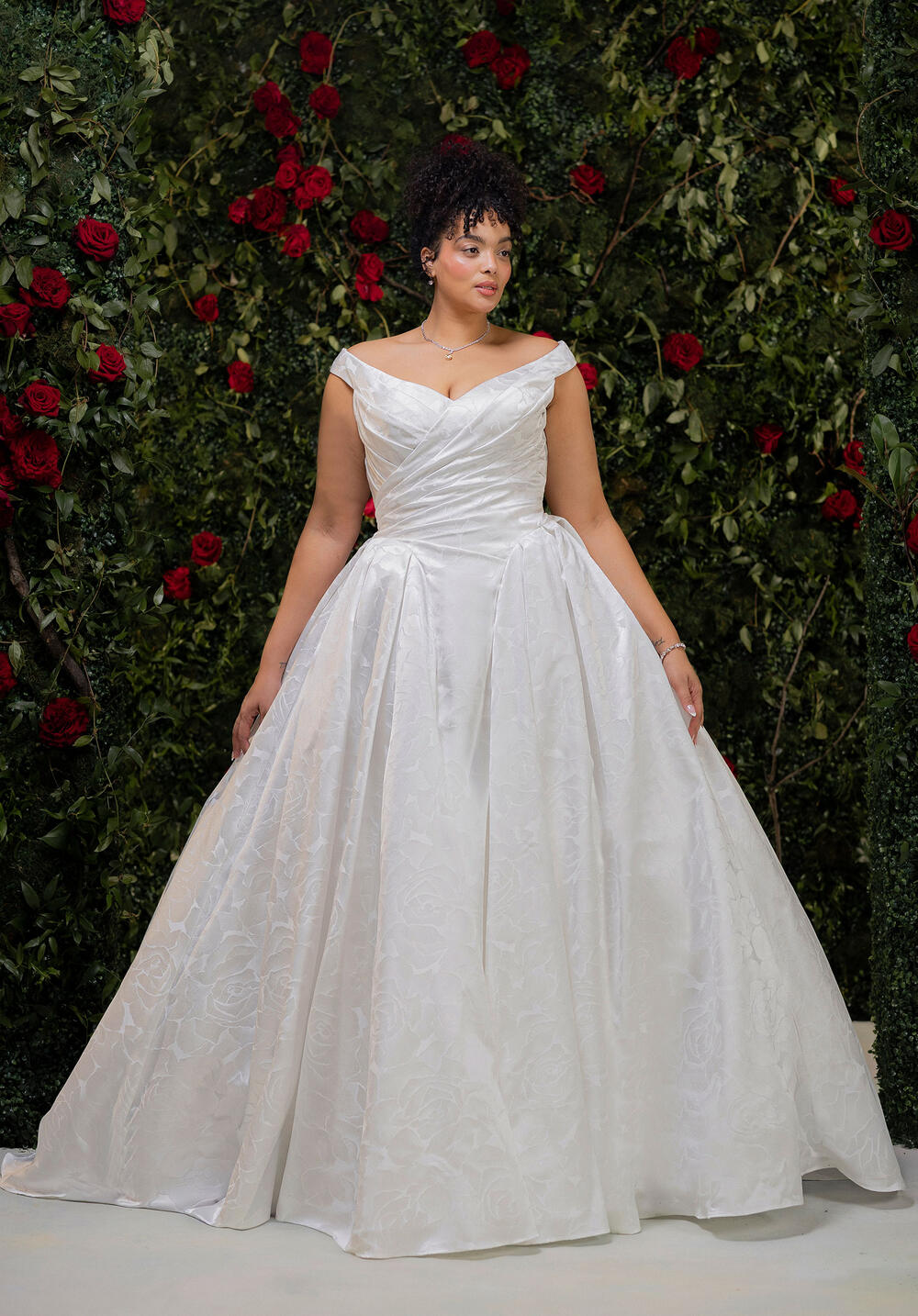 Morilee off-the-shoulder satin ball gown in front of a rose wall