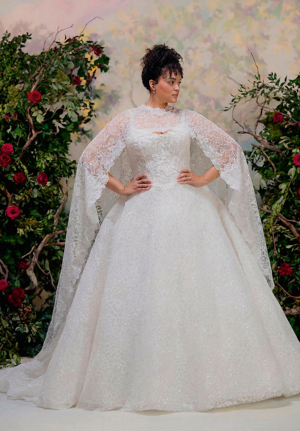 Morilee sparkling ball gown with a sheer lace cape in front of a rose wall