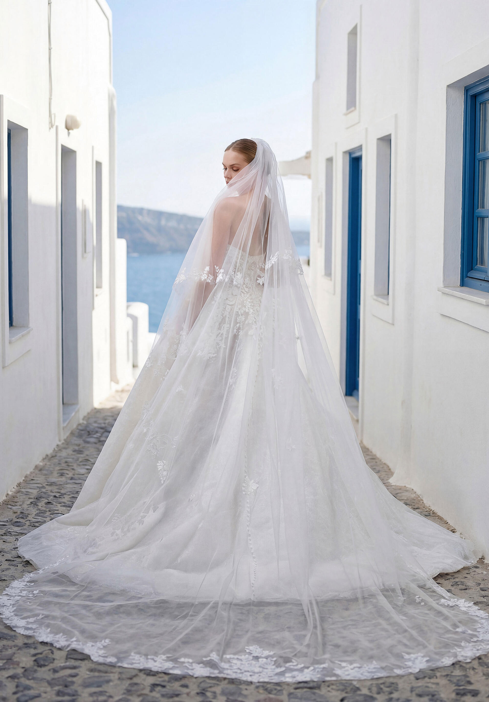 A bride walking away down a whitewashed lane, her Morilee gown's long lace train and veil trailing behind