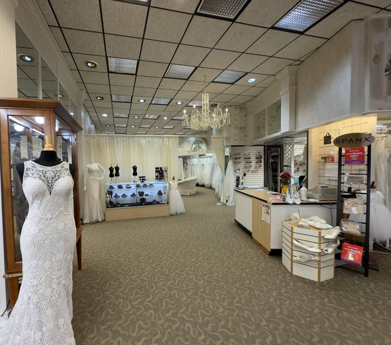 The sales floor, with a lace gown on a dress form, the jewelry case, a shoe display, and racks of wedding gowns under a chandelier