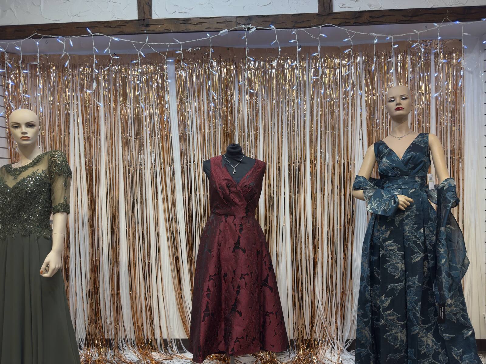 Three evening gowns on mannequins in front of a gold tinsel backdrop